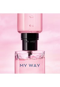 Nahaufnahme von rosa Flüssigkeit, die aus einem durchsichtigen Behälter in eine klare quadratische Parfümflasche mit der Aufschrift "MY WAY" gegen einen sanften rosa Hintergrund tropft.