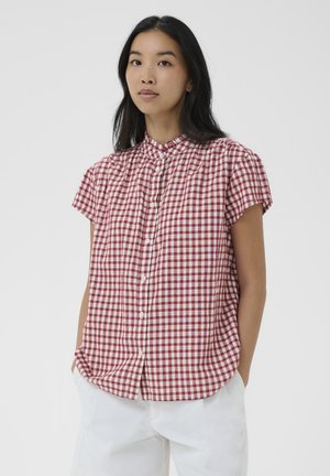 Femme aux longs cheveux noirs portant une chemise à manches courtes à carreaux rouges et blancs et un pantalon blanc, debout les mains dans les poches.