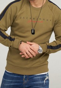 Hombre con sudadera oliva de Jack & Jones con rayas negras en las mangas, jeans ajustados azules desgastados, reloj de pulsera plateado y collar de pendant negro.