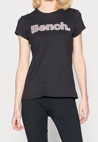 Camiseta de algodón negra con mangas cortas y cuello redondo. La palabra "Bench." está prominentemente exhibida en un diseño contrastante.