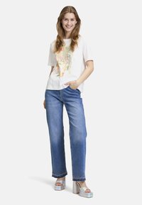 T-shirt blanc avec imprimé floral coloré, jeans larges bleus avec ourlet orné et sandales à plateforme en argent brillant.
