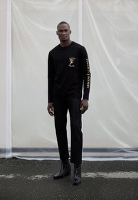 Melna garroku krekls ar zelta logotipu uz krūtīm un piedurknēm, kopā ar tumšām džinsa biksēm un melniem potīšu zābakiem ar papēžiem.