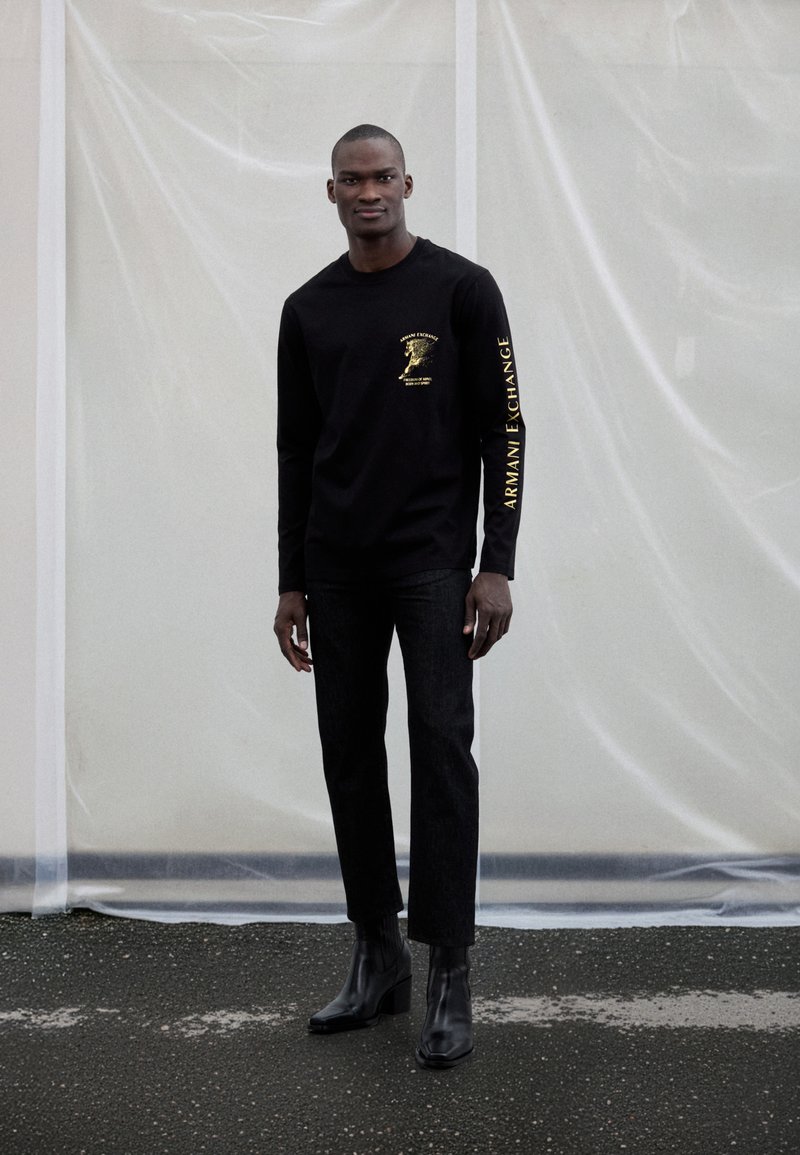 Melna garroku krekls ar zelta logotipu uz krūtīm un piedurknēm, kopā ar tumšām džinsa biksēm un melniem potīšu zābakiem ar papēžiem.