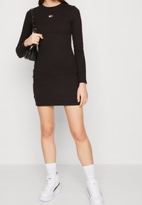 Black long-sleeve dress with a fitted silhouette, featuring a curved seam detail under a small colorful logo. Paired with white chunky sneakers.