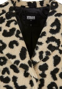 Bež kaput od lažnog krzna s crnim leopard uzorkom, s klasičnim ovratnikom i unutrašnjom oznakom. Mekana tekstura i oversized dizajn.