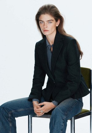 Jeune femme aux longs cheveux portant un blazer noir et un jean bleu, assise sur une chaise verte les mains jointes.