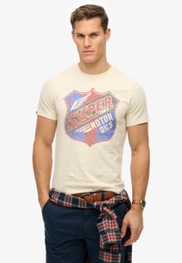 Camiseta de algodón color crema con un colorido diseño gráfico de un escudo que lleva el texto "SUPER DRY MOTOR OILS". Cintos y camisa de cuadros atada a la cintura.
