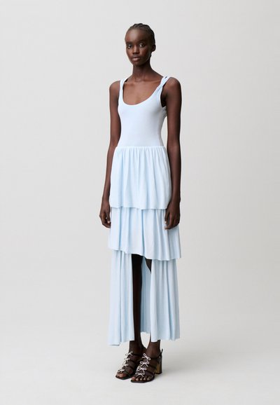 Donna alta con un vestito senza maniche azzurro chiaro a strati con scollatura ampia e sandali neri con tacco e lacci bianchi, in piedi su sfondo grigio.