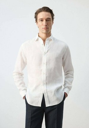 Hombre con camisa blanca de lino abotonada y pantalones oscuros con rayas finas, de pie con las manos en los bolsillos contra un fondo liso.