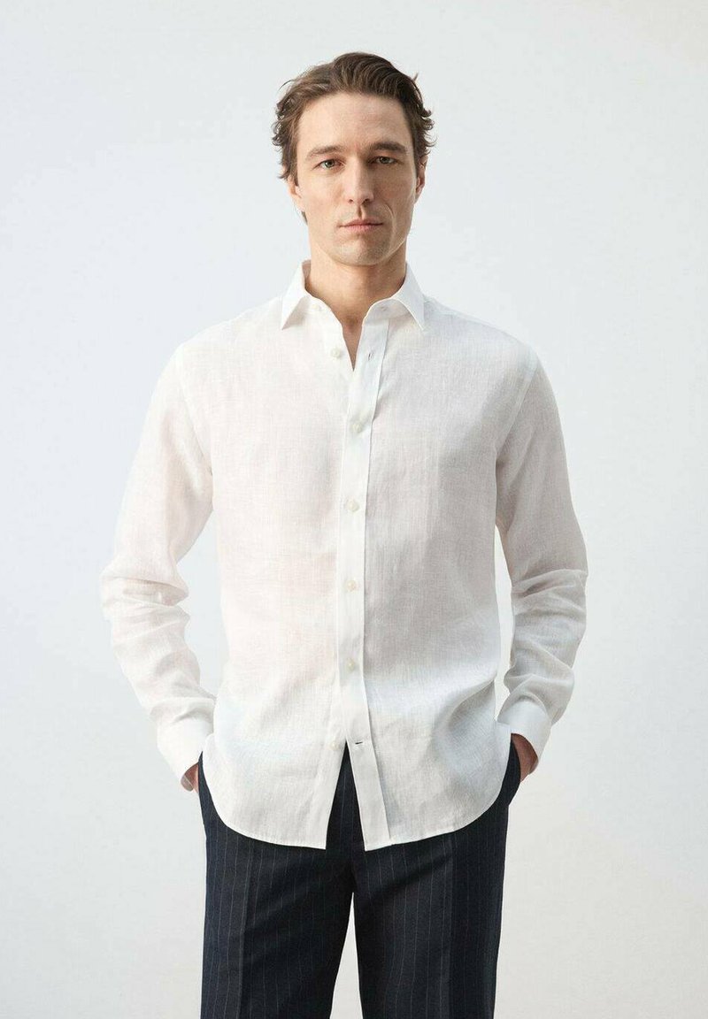 Homme portant une chemise blanche en lin avec boutons et un pantalon à fines rayures foncées, debout les mains dans les poches devant un fond uni.