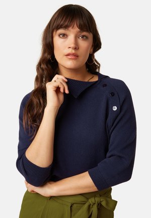 Vrouw met lang bruin haar die een marineblauwe trui draagt met knopen op de schouder en olijfgroene broek die in de taille gestrikt is.