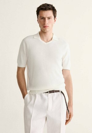 Giovane uomo che indossa una polo a maniche corte in maglia bianca infilata in pantaloni bianchi con una cintura intrecciata marrone, in piedi con una mano in tasca.