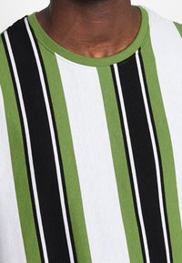 Zielona, czarna i biała koszulka w paski z okrągłym dekoltem. Materiał wydaje się miękki, z wyraźnymi naprzemiennymi wzorami kolorystycznymi.