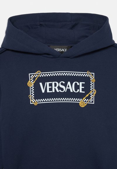 Sudadera con capucha azul marino que presenta un logo blanco con un borde a cuadros y alfileres de seguridad dorados como acentos. Tejido suave, diseño casual.