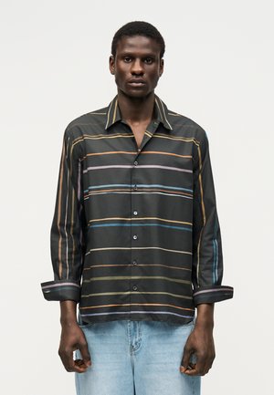 Homme portant une chemise noire boutonnée avec des rayures horizontales et verticales multicolores, associée à un jean bleu clair.