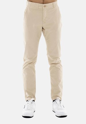 Pantaloni beige slim-fit indossati da una persona sopra sneakers bianche con suola nera, in piedi su uno sfondo chiaro e uniforme.