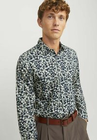 Chemise à boutons florale avec fond marine, ornée de petites fleurs blanches et bleu clair. Associée à une ceinture en cuir marron et à un pantalon plissé.