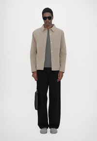 Light brown jacket with snap buttons and two chest pockets, gray T-shirt underneath, black wide-leg pants, gray shoes, and black sunglasses.