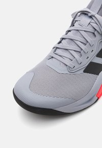 Light grey athletic shoe with a textured mesh upper, black accents, flat laces, and a red outsole detail. Rounded toe and cushioned design.