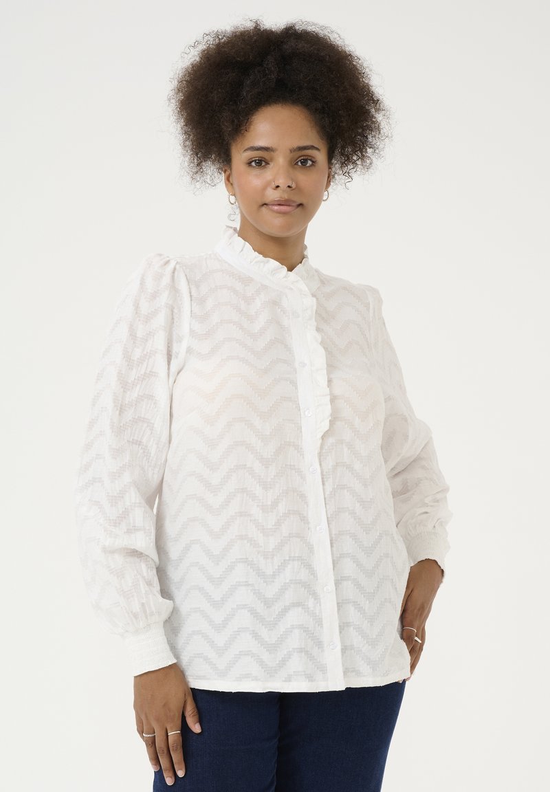 Blusa blanca con cuello volante, frente abotonado y patrón de textura en zigzag. Mangas largas con puños elásticos, combinada con jeans azul oscuro.