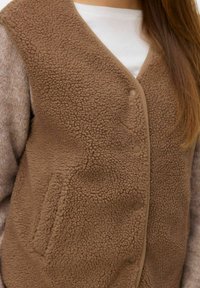 Brown fleece vest with button closure, soft texture, rounded neckline, and side pocket. Layered over a light-colored top.