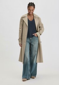 Beige trenchcoat met een dubbelbreasted ontwerp, brede revers en riemmanchetten, gecombineerd met een marineblauwe gebreide trui en een wijdvallende blauwe jeans.