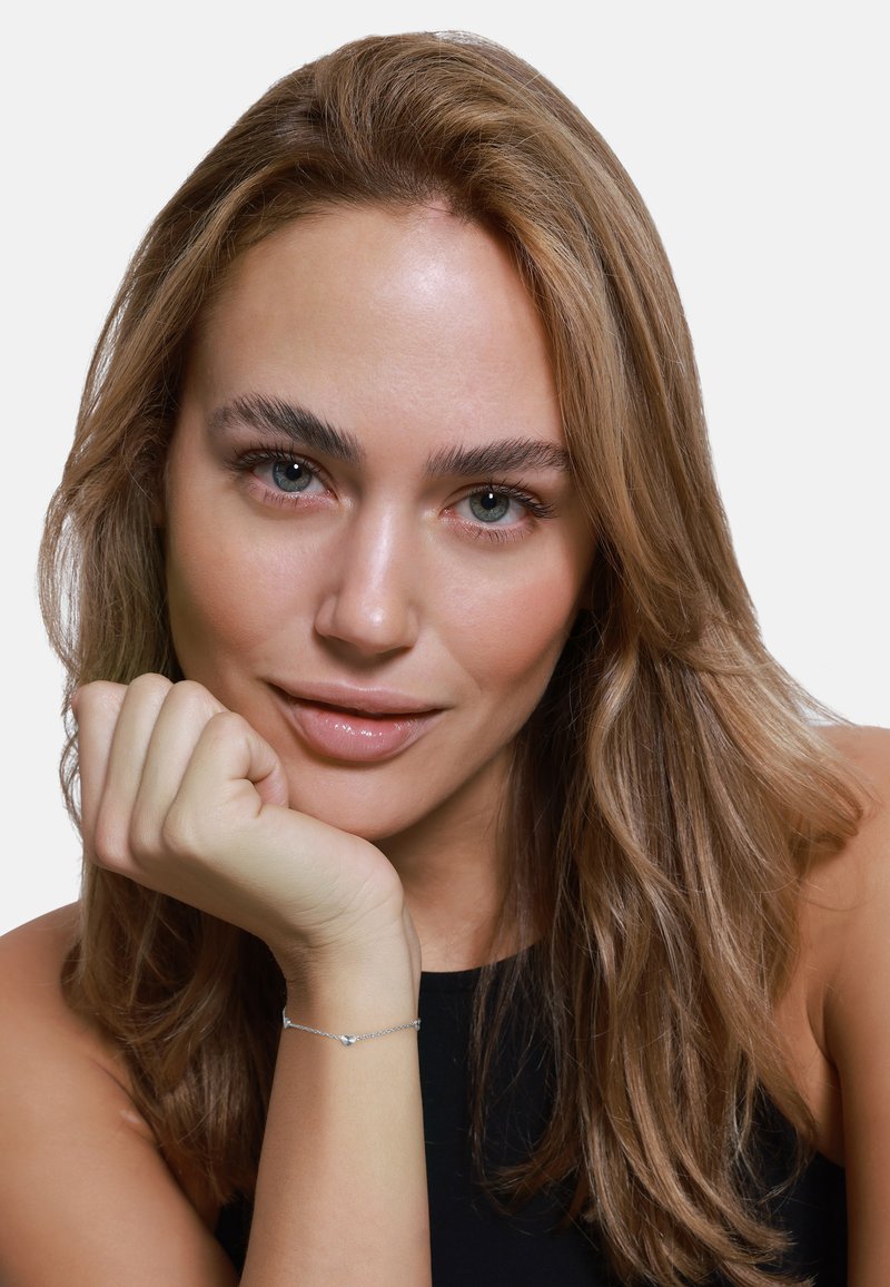 Silver bracelet with small heart accents, worn on a hand resting near the chin, featuring smooth skin and long, wavy hair.