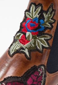 Botte en cuir marron avec des patchs floraux brodés en rouge, bleu, vert et noir près de la cheville et un panneau élastique latéral.