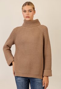 Maglione beige lavorato a maglia con colletto alto e arrotolato, caratterizzato da una texture a costine, maniche ampie e una vestibilità rilassata. Abbinato a jeans blu.