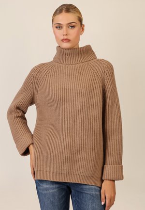 Beiger Strickpullover mit hohem, gerolltem Kragen und gerippter Textur, mit weiten Ärmeln und lockerer Passform. Kombiniert mit blauen Jeans.