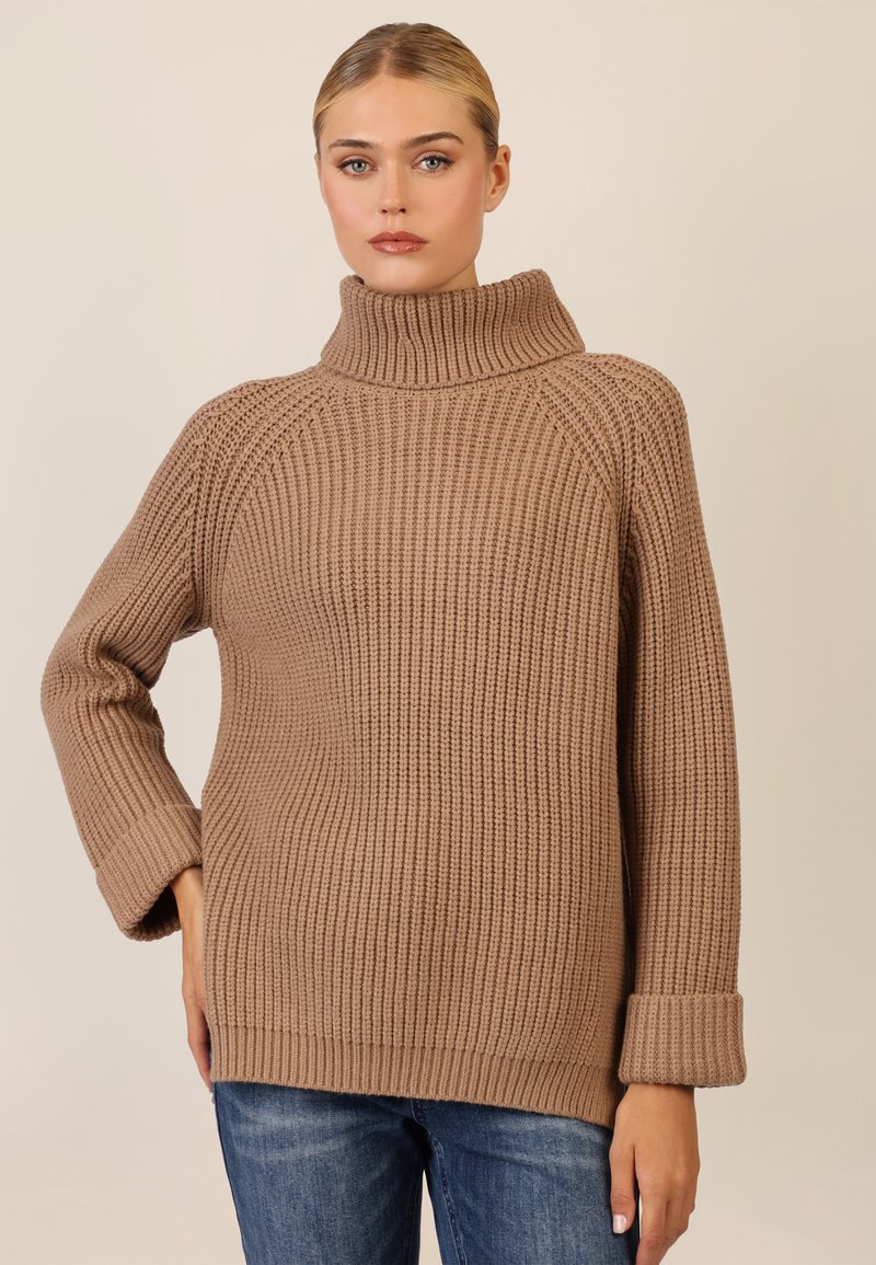 Beige gebreide trui met een hoge, omgeslagen kraag en geribbelde textuur, met brede mouwen en een relaxte pasvorm. Gekleed met blauwe jeans.