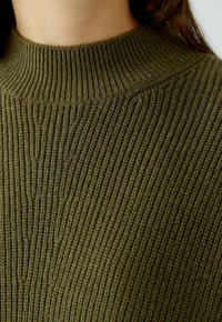 Olivgrüner Strickpullover mit geripptem Muster und hohem Kragen. Eine Nahaufnahme hebt die Weichheit des Materials und die detaillierte Naht hervor.
