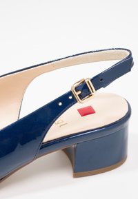 Sandale à talon bas en cuir verni bleu foncé avec bride à boucle dorée et semelle intérieure beige coussinée dotée d'un petit détail carré rouge.