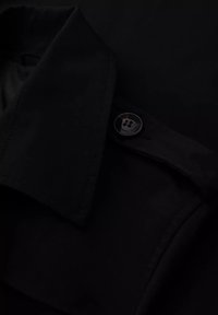 Czarna tkanina kurtki z kołnierzem, z okrągłym guzikiem z logo. Gładka tekstura i dopasowany krój.