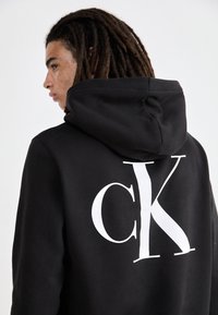 Sudadera negra con capucha que presenta un gran logo blanco "ck" en la parte posterior. Tejido suave con un corte relajado y capucha ajustable.