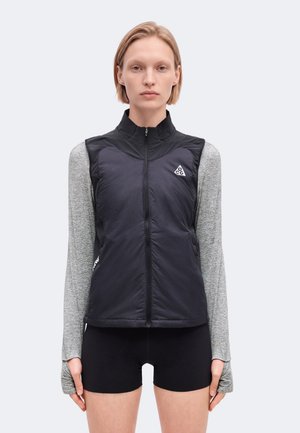 Un mannequin se tient face à l’avant, portant un gilet noir zippé par-dessus un haut gris à manches longues et un short noir.