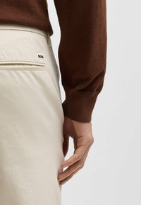 Beige pants with a back pocket featuring a black "BOSS" logo, paired with a brown textured sweater, showing the arm and hand.