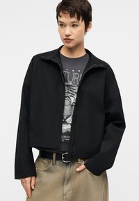 Jaunas žmogus su trumpais tamsiais plaukais dėvi juodą švarkelį ant tigro piešinio marškinėlių ir išblukusias smėlio spalvos džinsines kelnes su juodu diržu.