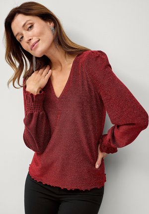 Rode glanzende blouse met een V-hals, lange pofmouwen en gerimpelde manchetten. Zachte textuur met een gegolfde zoom. G gedragen met zwarte broeken.