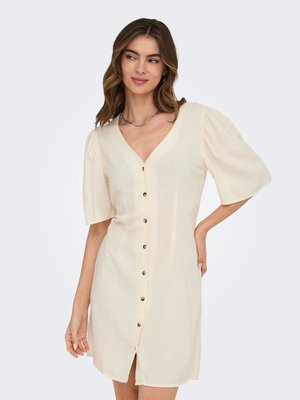 Femme portant une robe crème boutonnée avec des manches bouffantes jusqu'au coude, debout avec une main sur la hanche, regardant sur le côté.