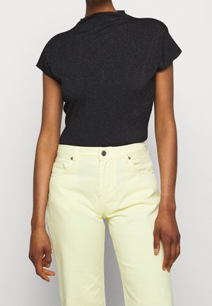 Osoba ubrana w czarny dopasowany top z krótkim rękawem i jasnożółte spodnie z wysokim stanem, stojąca na jednolitym tle.