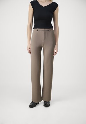 Femme portant un haut noir à manches courtes, un pantalon large couleur taupe et des chaussures de ville noires, debout sur un fond clair uni.