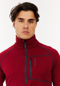 Crvena polar jakna na zip s visokim ovratnikom, teksturiranim materijalom, kontrastnim sivim šavovima i džepom na prsima s zipom. Sadrži logotip.
