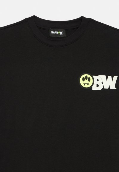 Camiseta negra con cuello redondo, que presenta un pequeño logotipo blanco "BW" y una cara sonriente amarilla con colmillos en el pecho.