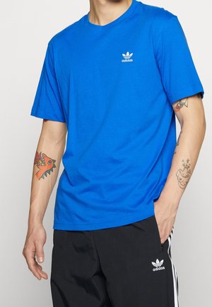 T-shirt en coton bleu à manches courtes, col ras du cou, avec un petit logo blanc Adidas sur la poitrine. Porté avec un pantalon noir à bandes blanches.