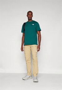 Turkizen bombažna majica s kratkimi rokavi, kombinirana s kameljnimi cargo hlačami z stranskimi žepi. Čevlji so svetlo sivi z teksturiranimi detajli.