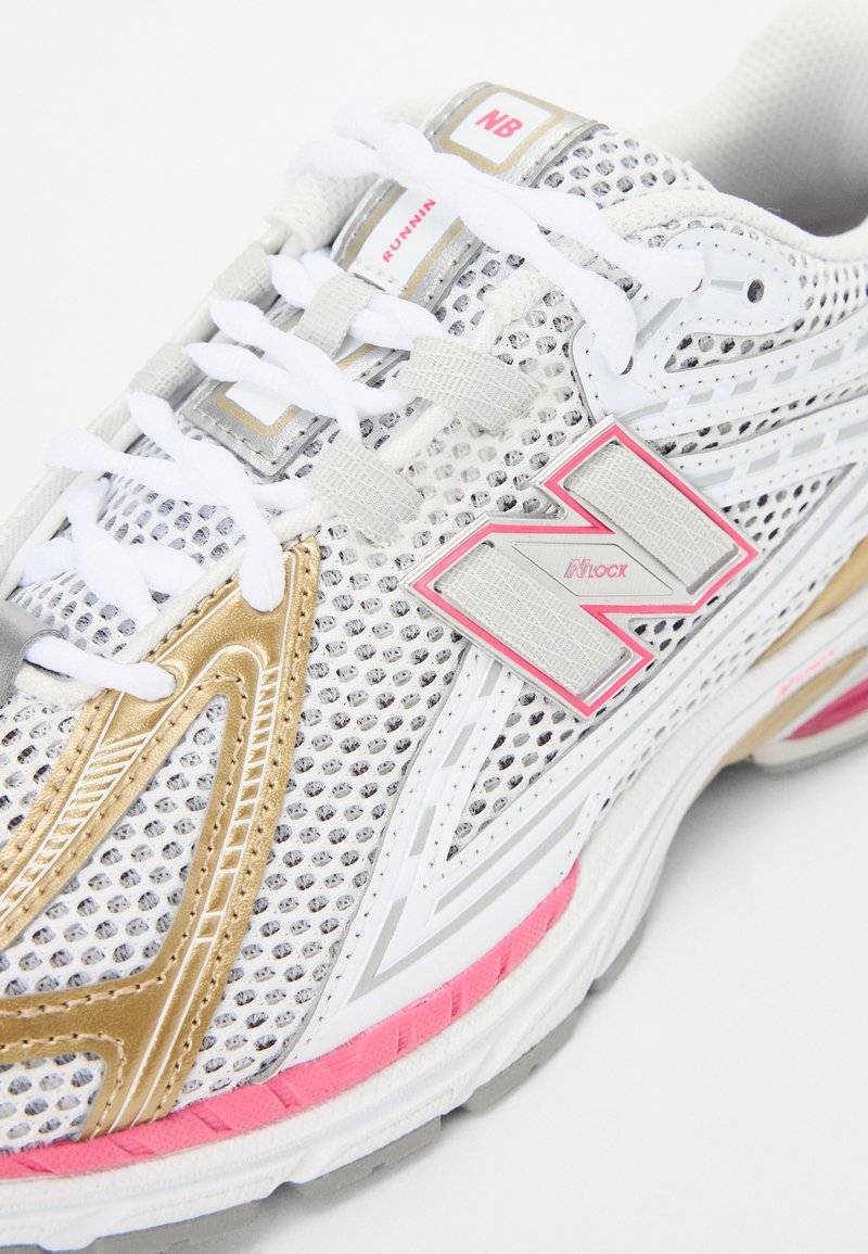 Zapatilla de running blanca con diseño de malla, acentos en dorado y rosa, con herrajes grises, cordones gruesos blancos y una suela con patrón.