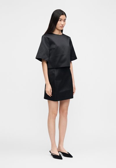 Melna crop top ar īsām piedurknēm, kombinēts ar melnu cieši piegulošu mini svārku, abi no gludas auduma. Melni mulei ar ieapaļu ēsas galiņiem papildina izskatu.