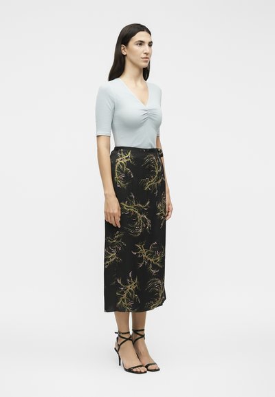 Γυναίκα με μακριά σκούρα μαλλιά φοράει ένα γαλάζιο ρουχαριστό τοπ, μαύρη φλοράλ μίντι φούστα και μαύρα ψηλοτάκουνα σανδάλια με λουράκια.