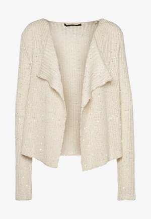 Femme portant un cardigan en tricot crème, un haut noir, un jean skinny foncé avec revers, des bottines noires, tenant une pochette noire.